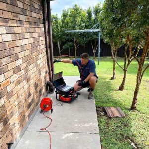 Plumber operating a Razorback drain camera inserted into a pipe beside a house